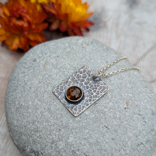 Silver Rectangle Necklace with Amber resting on a smooth grey stone, with orange flowers softly blurred in the background.