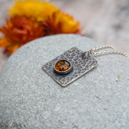 Angled view of the Silver Rectangle Necklace with Amber lying on a grey stone, showing the warm amber glow against the oxidised hammered silver.