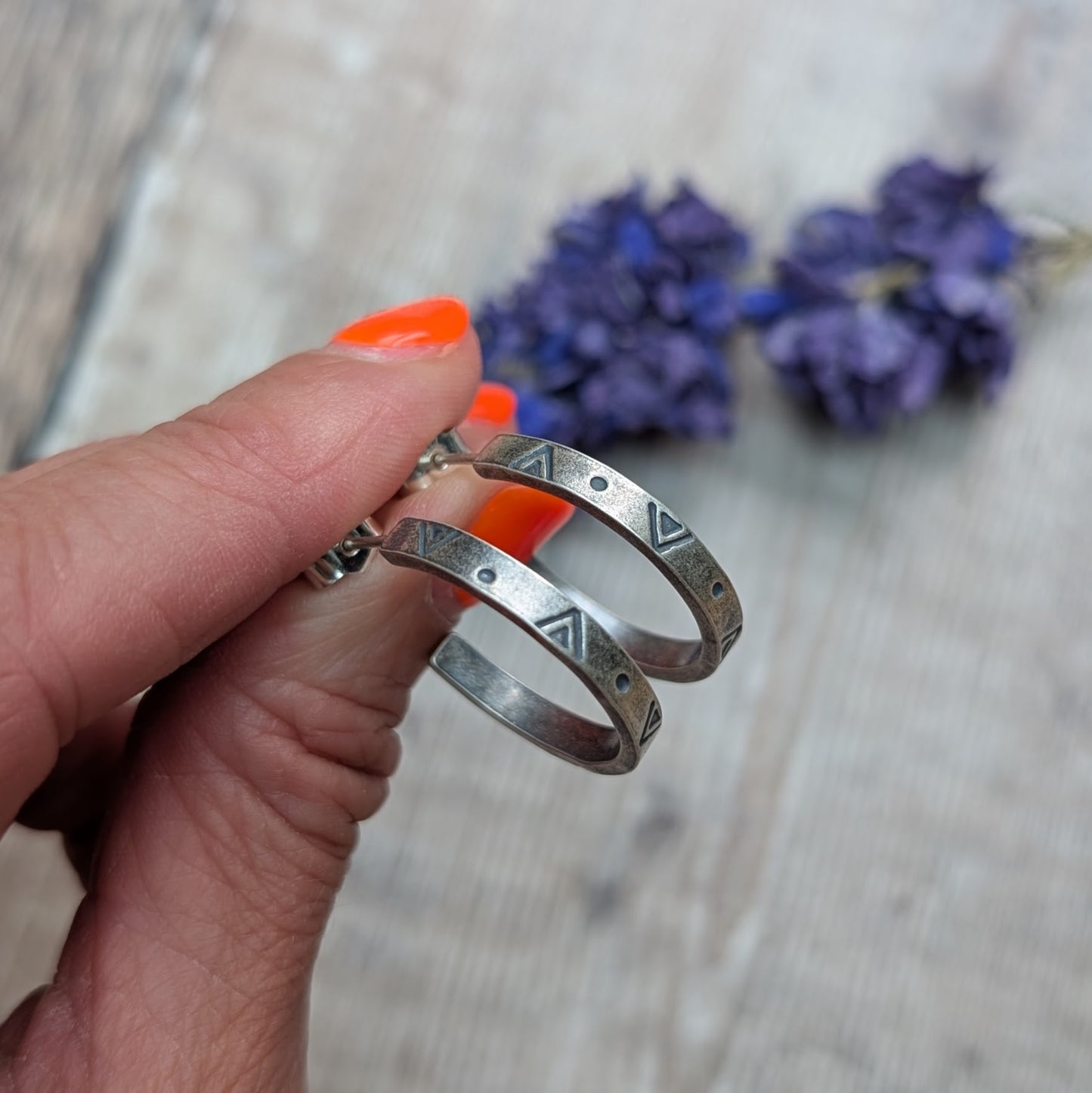 Close-up of a hand holding the silver geometric hoop earrings, with a backdrop of blurred purple flowers and wooden surface.