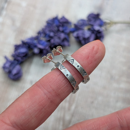 Silver geometric hoop earrings balanced across a finger, showing their hand-stamped triangle and dot pattern.