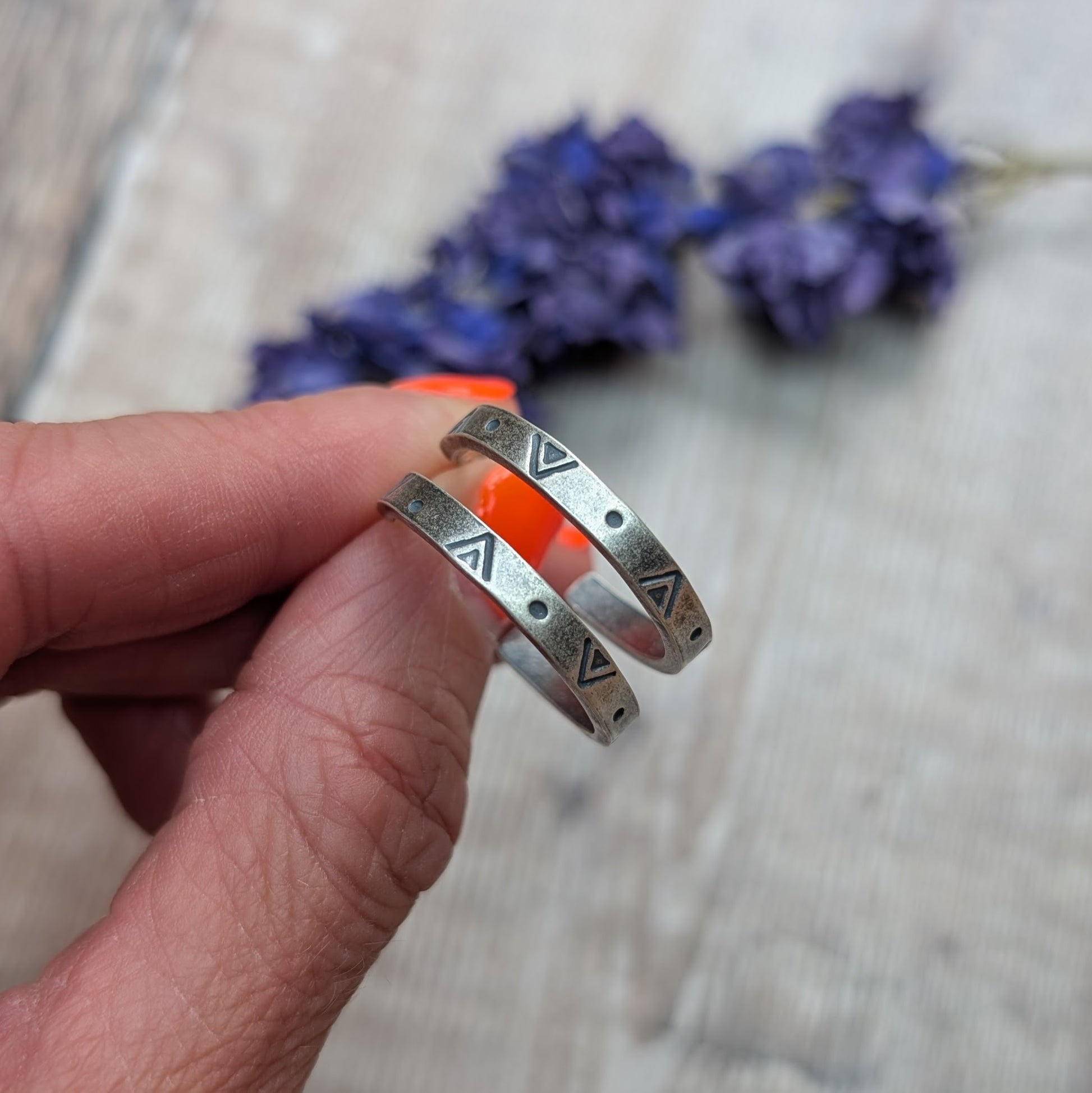 Close-up of a hand holding the silver geometric hoop earrings, with a backdrop of blurred purple flowers and wooden surface.