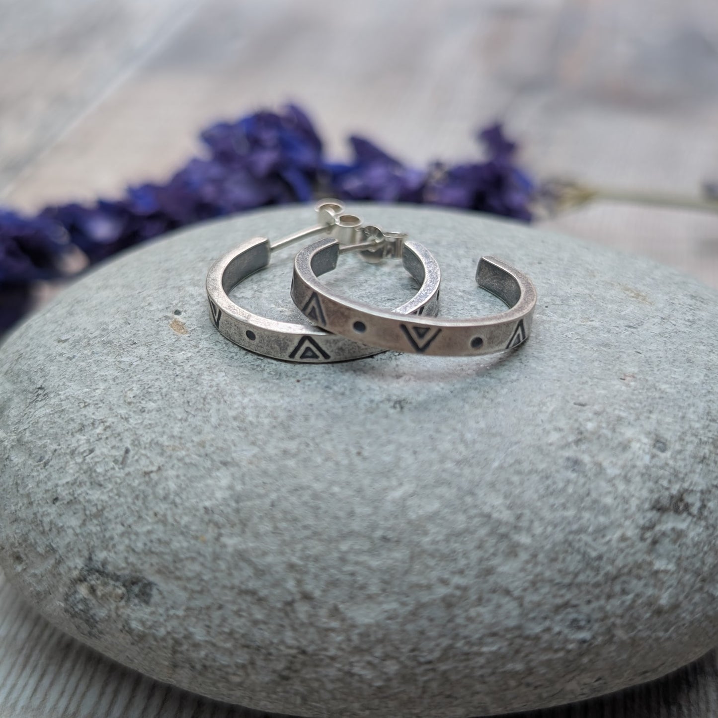 A pair of silver geometric hoop earrings resting on a smooth grey stone, with dried purple flowers in the background.