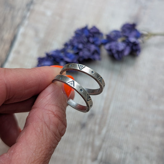Close-up of a hand holding the silver geometric hoop earrings, with a backdrop of blurred purple flowers and wooden surface.