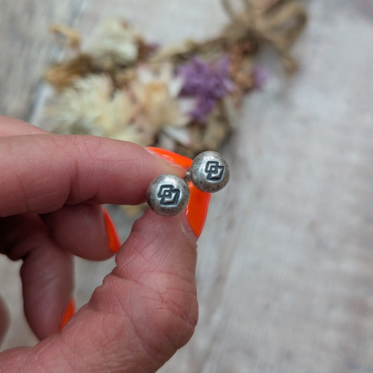Close-up of a hand holding a pair of Silver Geometric Pebble Studs in front of a softly blurred background with dried flowers.