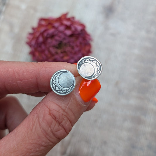 Pair of handmade silver large moon disc stud earrings held between fingers, showing engraved crescent moon design.