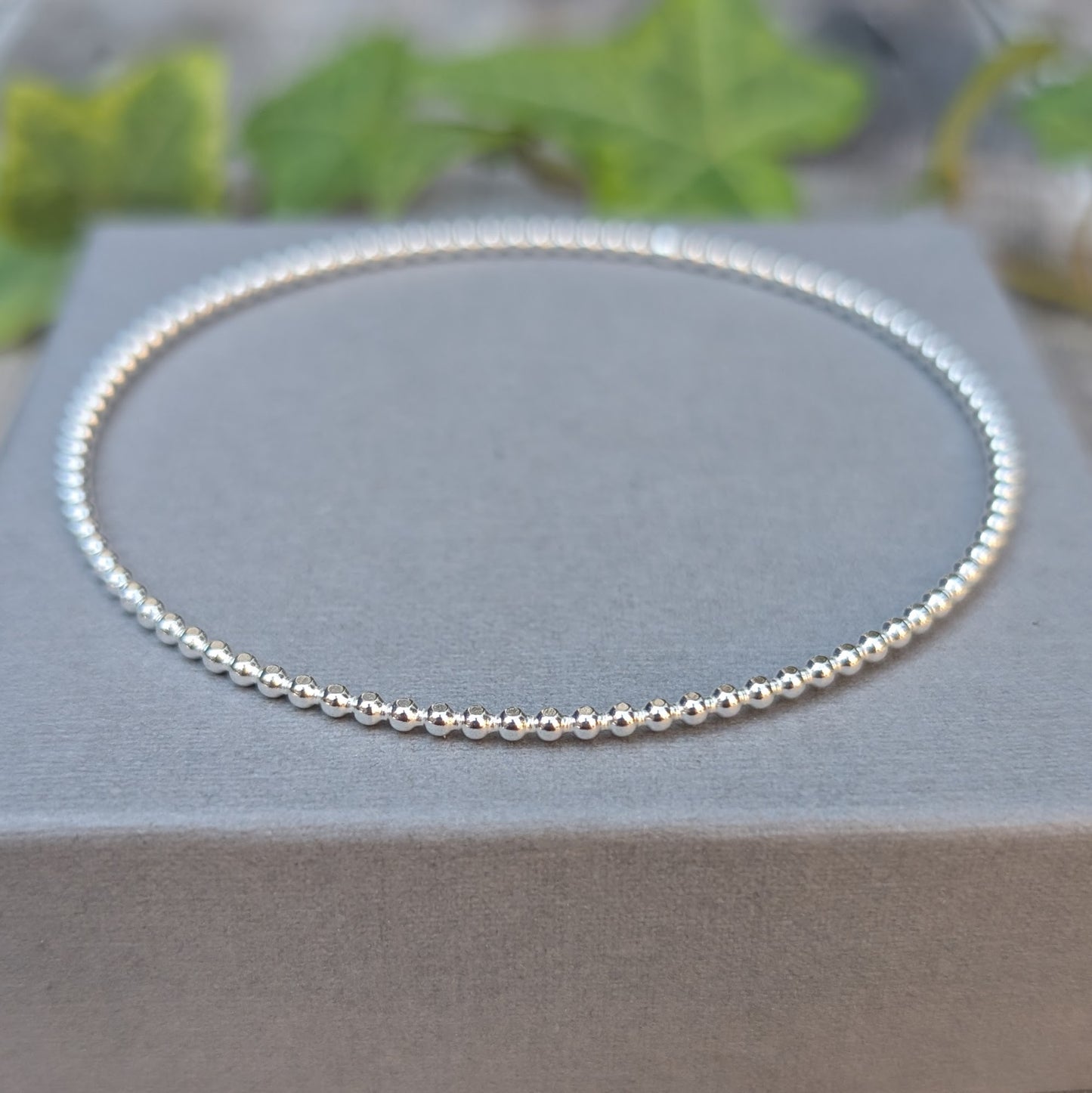 Silver 2mm Beaded Round Bangle handmade from recycled Sterling Silver, displayed on a grey jewellery box with green leaves in the background.