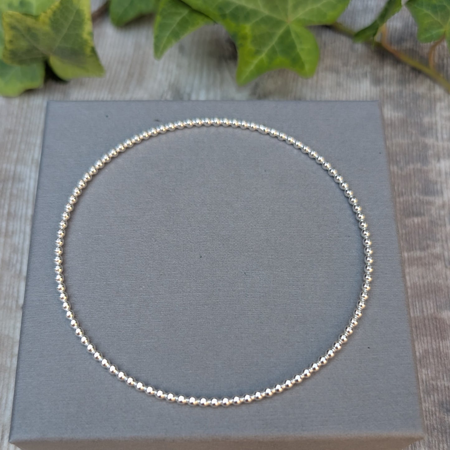 Silver 2mm Beaded Round Bangle handmade from recycled Sterling Silver, shown flat on a grey jewellery box.