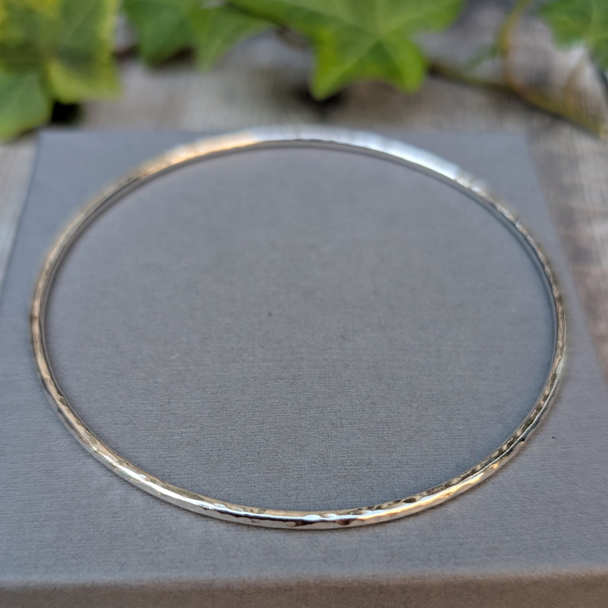 Silver 2mm Hammered Round Bangle handmade from recycled Sterling Silver, displayed flat on a grey jewellery box with green ivy in the background.