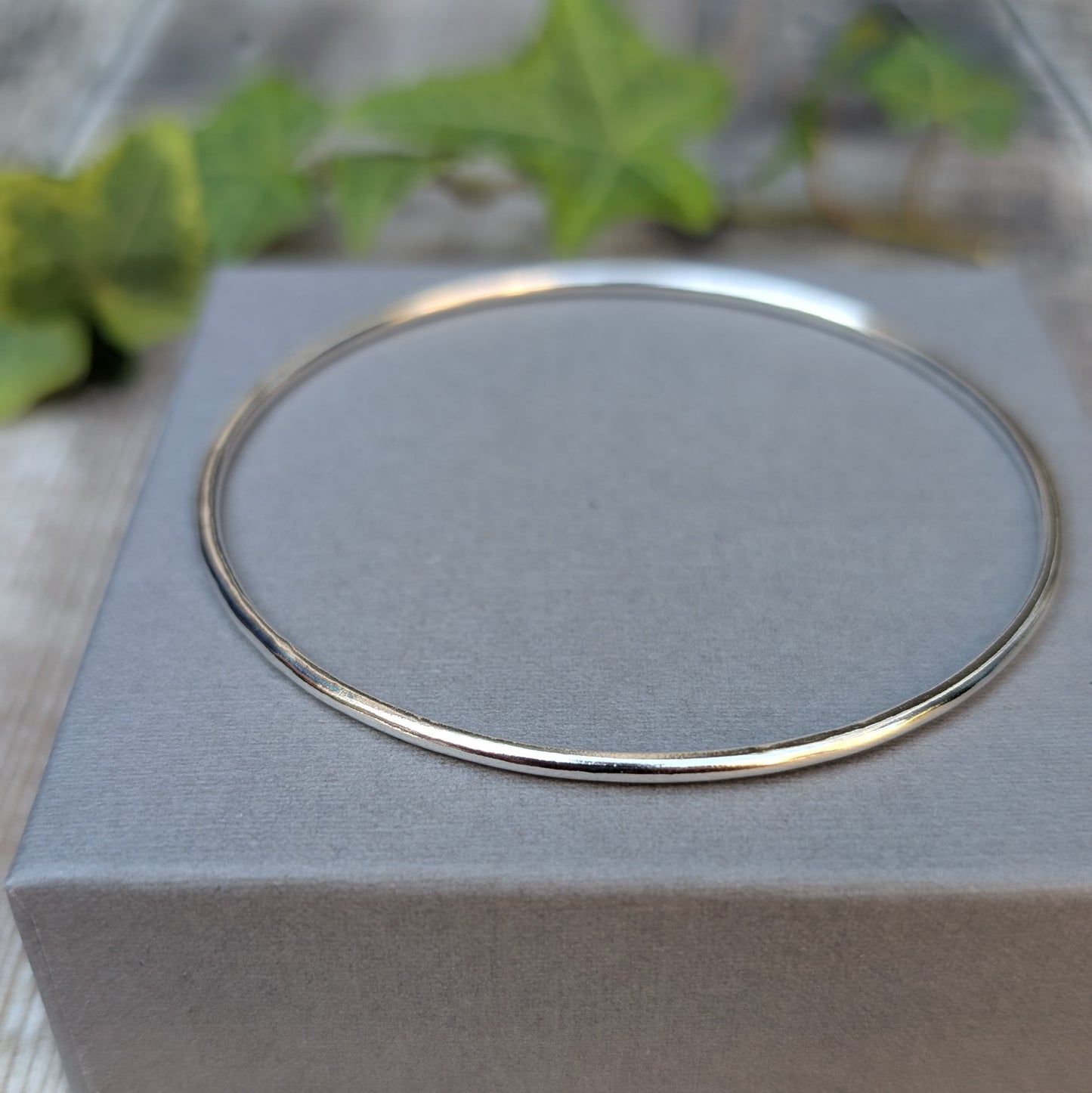 Silver 2mm Smooth Round Bangle displayed on a grey jewellery box, with a close up view of the simple smooth silver design.