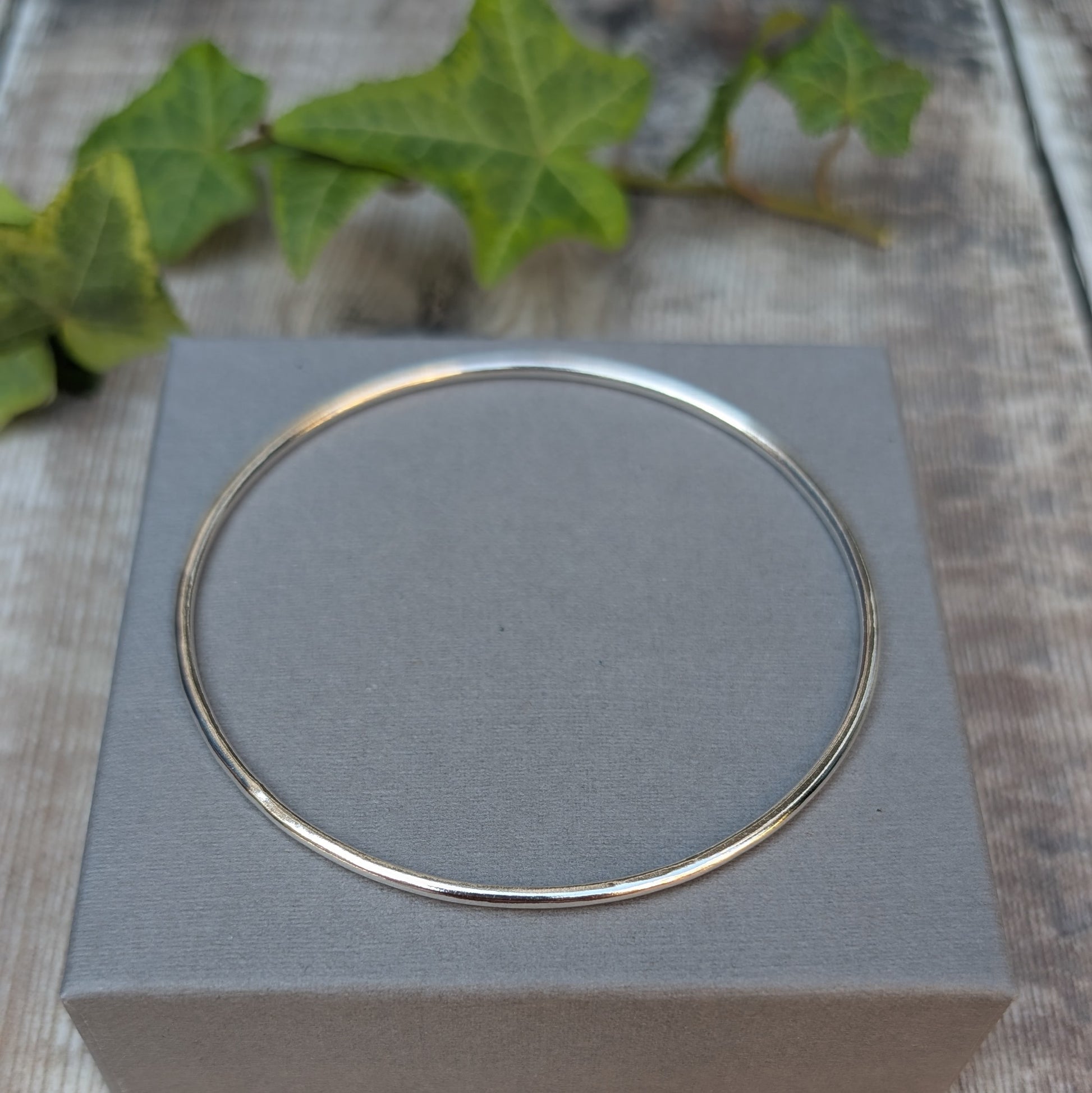 Silver 2mm Smooth Round Bangle handmade from recycled Sterling Silver, displayed on a grey jewellery box with green ivy in the background.