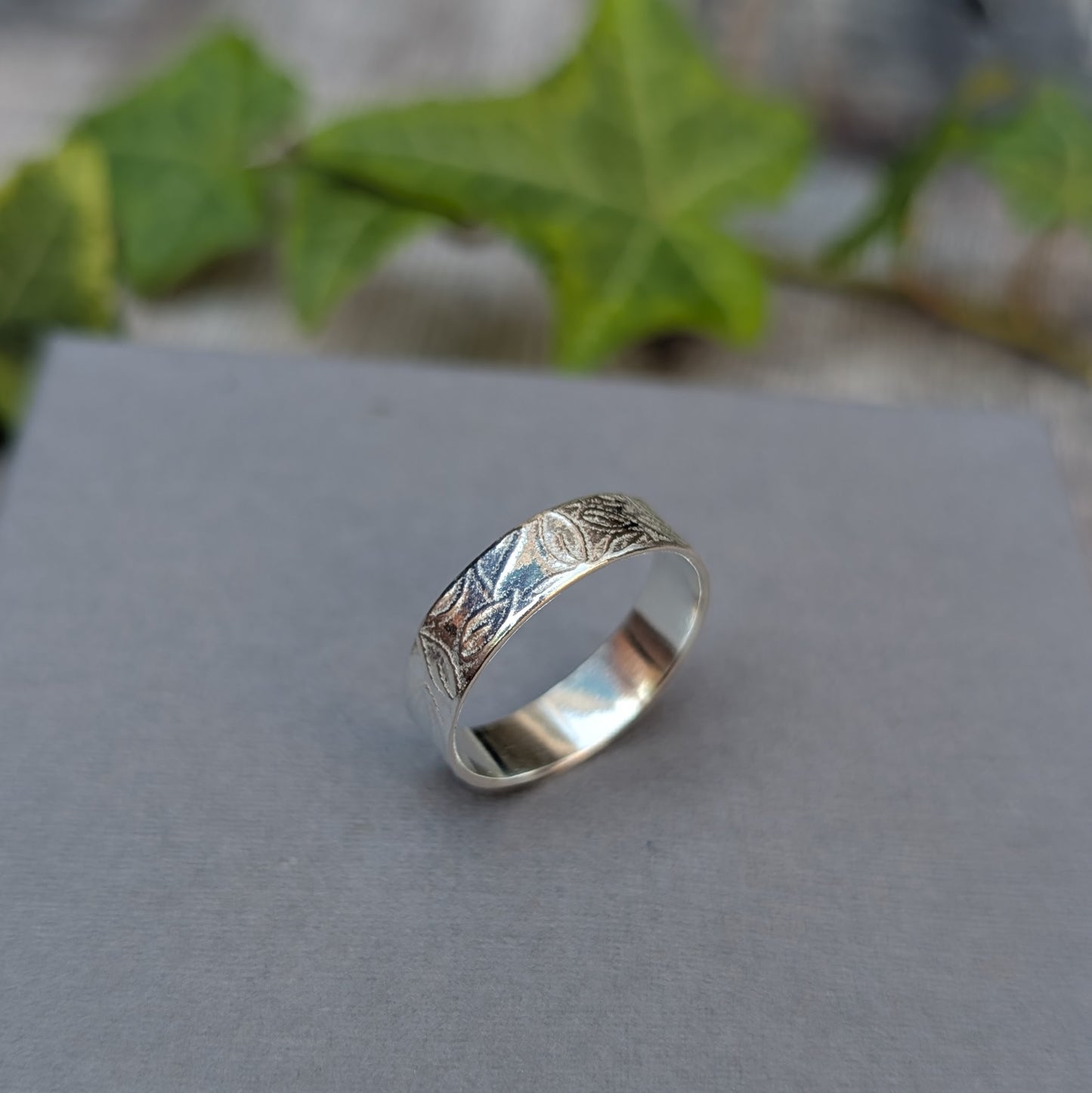 Silver 5mm Leaf Patterned Ring standing on a grey background with green leaves behind, showing the hand stamped leaf design around the band.