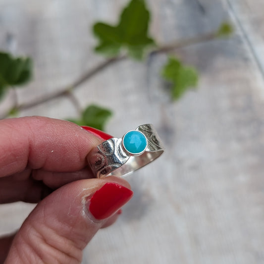 Close up of a silver oval patterned Amazonite wide band ring held in a hand, showing the bright blue Amazonite stone and detailed pattern on the wide silver band.