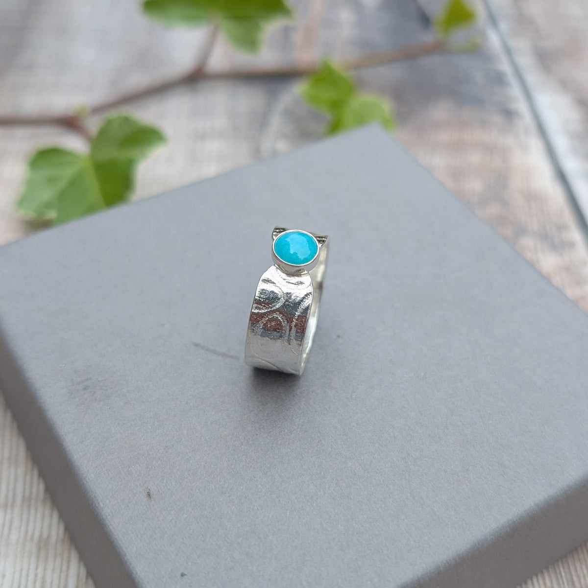 Silver oval patterned Amazonite wide band ring standing on a grey jewellery box, showing the blue Amazonite stone and patterned recycled Sterling Silver band.