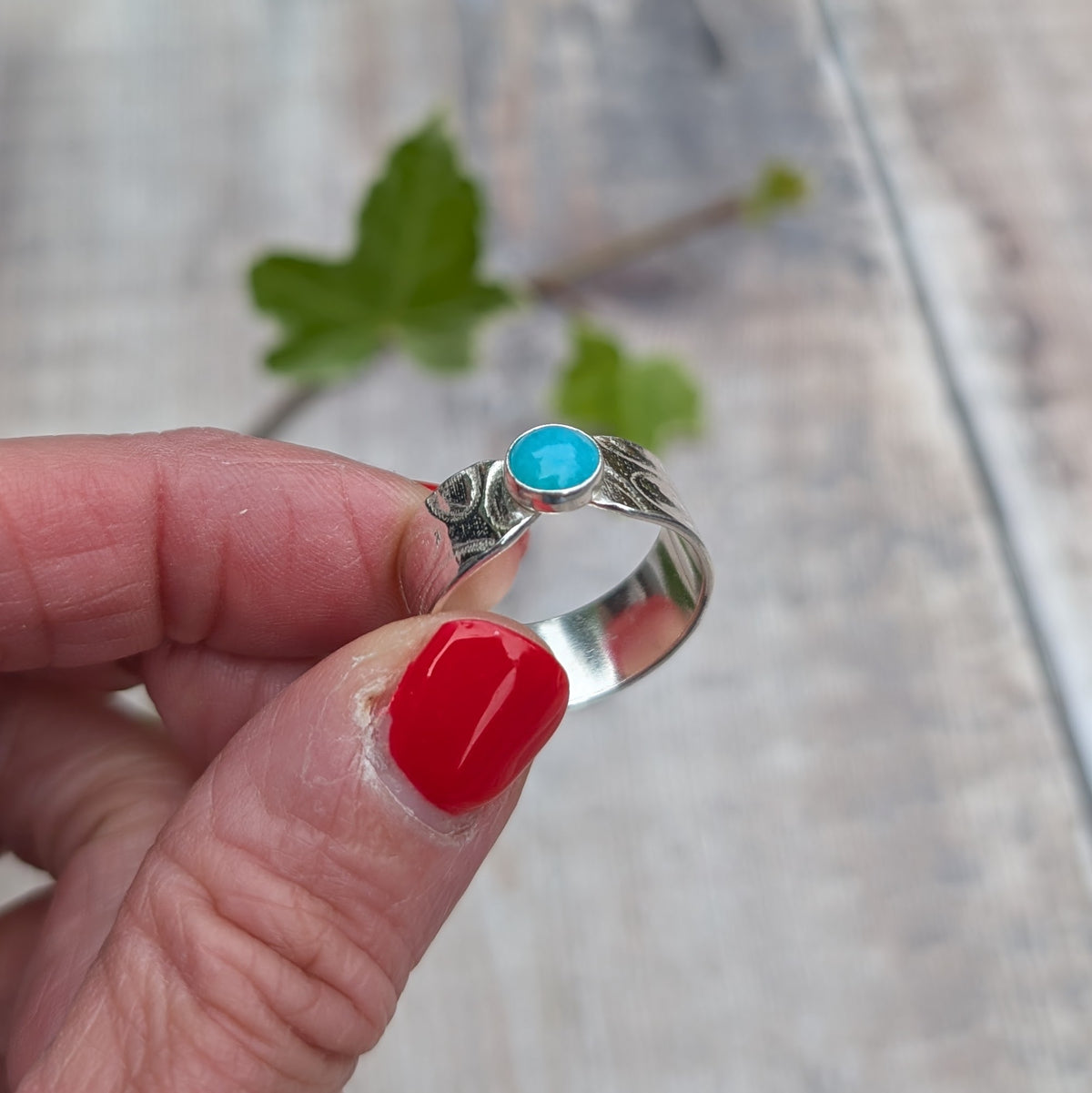 Close up of a silver oval patterned Amazonite wide band ring held between fingers, showing the blue Amazonite stone and patterned Sterling Silver band.