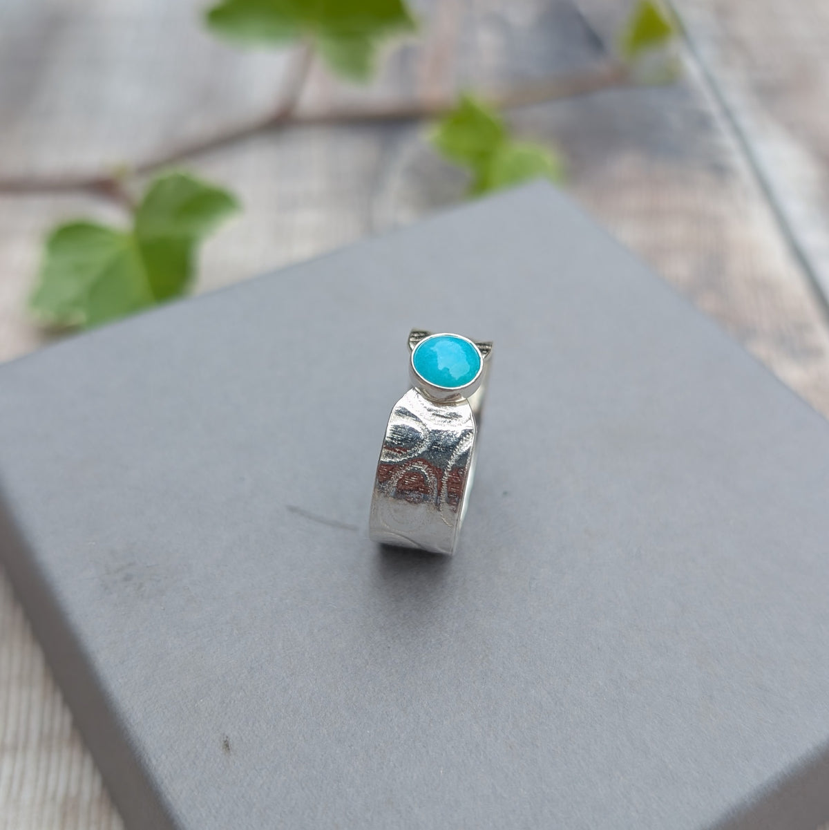 Silver oval patterned Amazonite wide band ring displayed upright on a grey jewellery box with the oval blue stone at the centre.