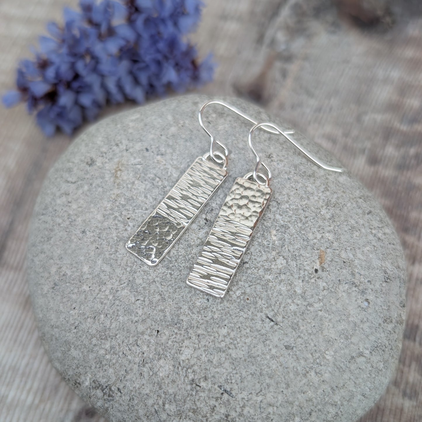 Silver asymmetrical hammered rectangle earrings with mixed textures, photographed on a smooth stone with dried flowers in the background