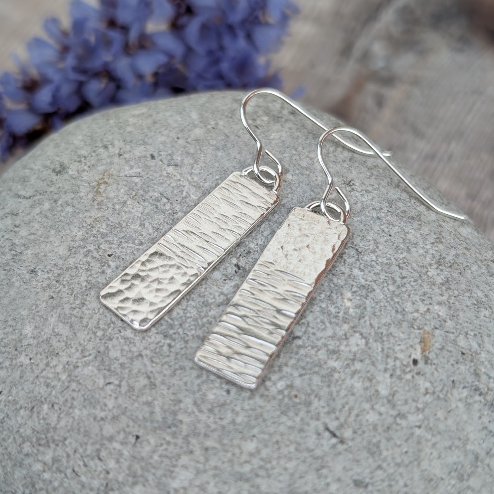 Silver asymmetrical hammered rectangle earrings showing contrasting linear and dimpled hammer marks on a stone surface
