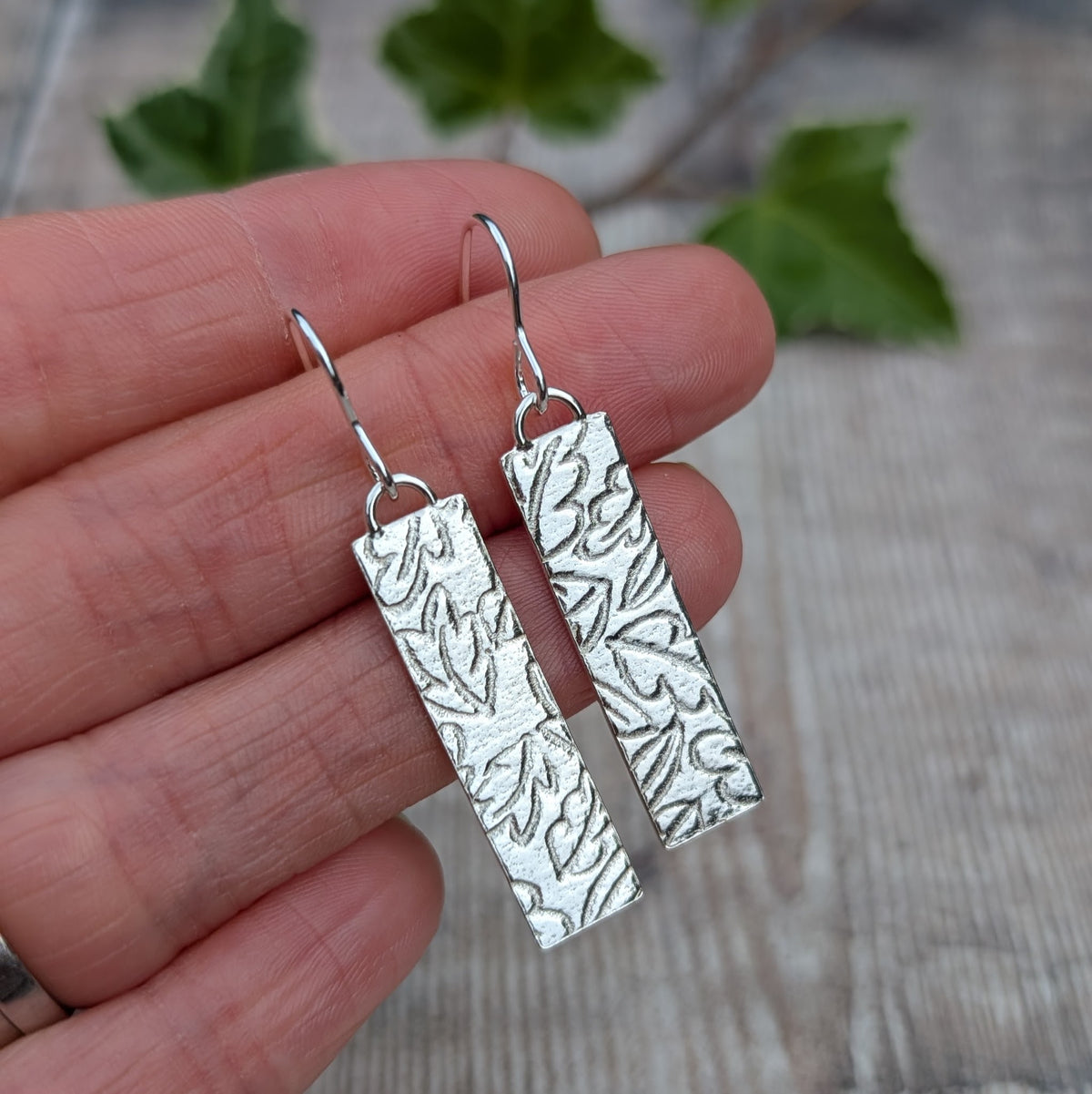 Close up of silver autumn leaves long rectangle earrings held in a hand, showing the detailed autumn leaf pattern created using a rolling mill.