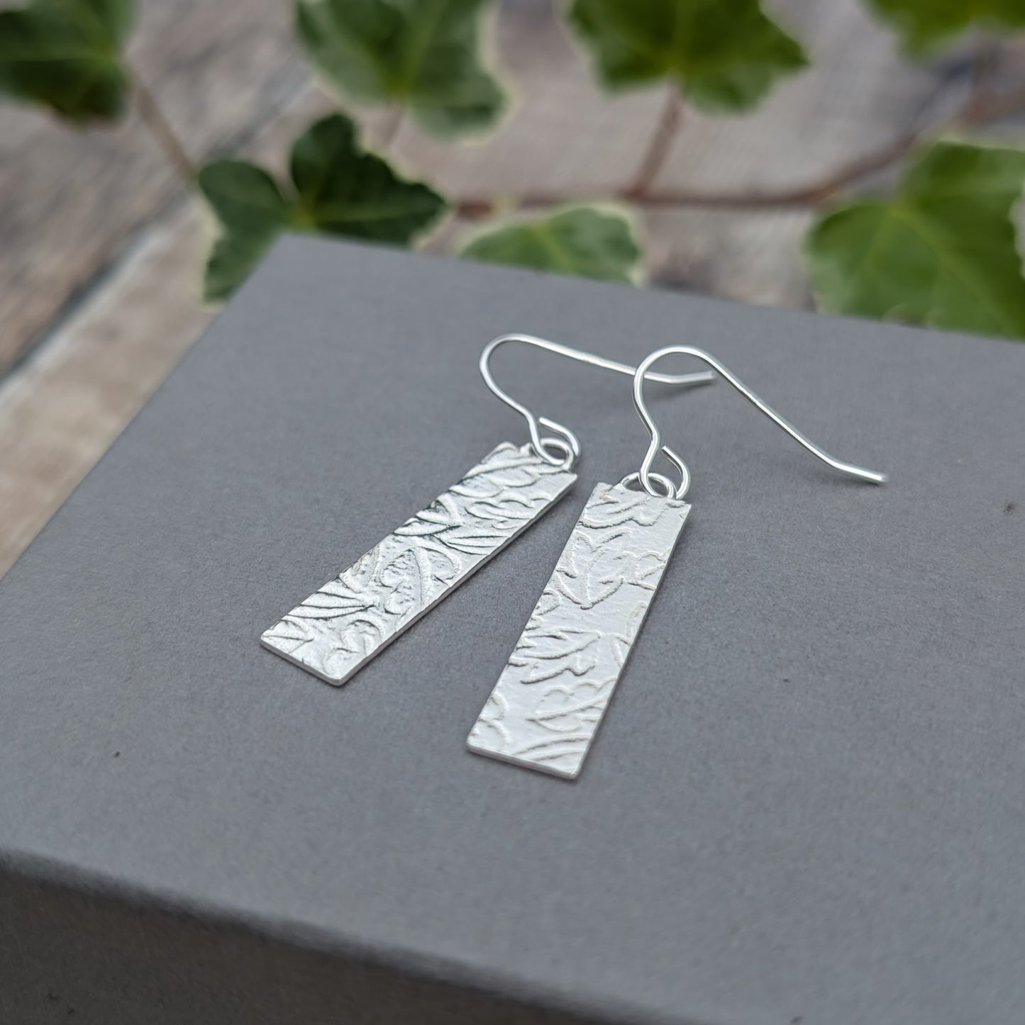Silver autumn leaves long rectangle earrings resting on a grey jewellery box, showing the textured leaf pattern and silver ear wires.