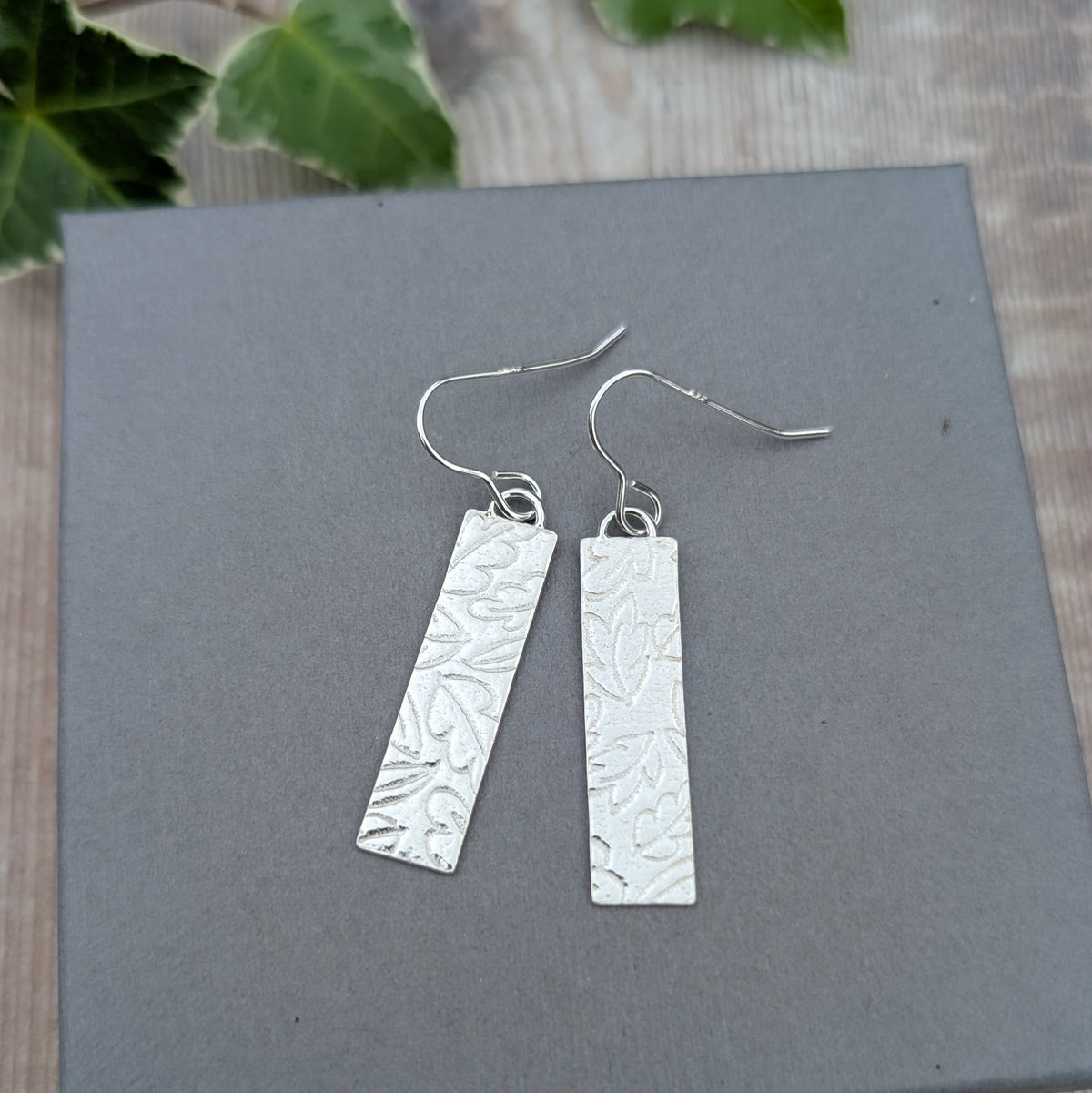 Front view of silver autumn leaves long rectangle earrings displayed on a grey jewellery box with a leafy background.
