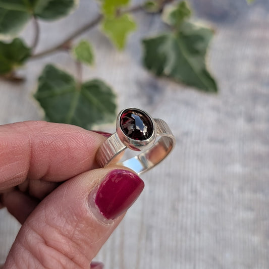 Close up of a silver bark textured garnet ring held between fingers, showing the polished oval garnet setting and bark inspired hammered silver band.
