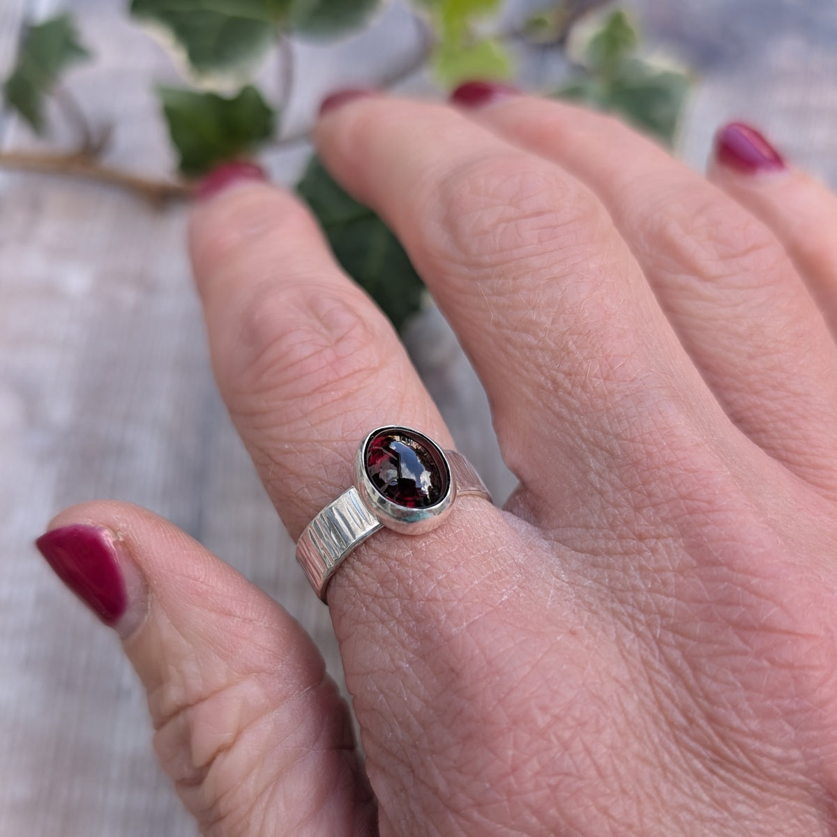 Silver bark textured garnet ring worn on a finger, showing the oval dark red garnet gemstone set in polished silver and the textured band.