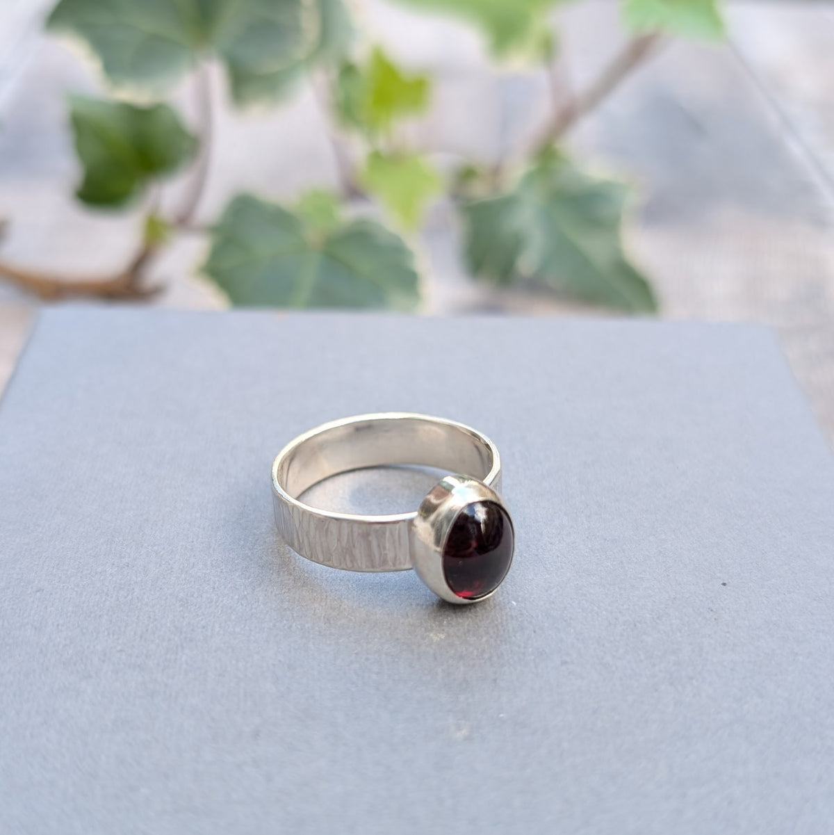 Handmade silver bark textured garnet ring resting on a grey surface, showing the wide textured sterling silver band and oval garnet gemstone in its silver setting.