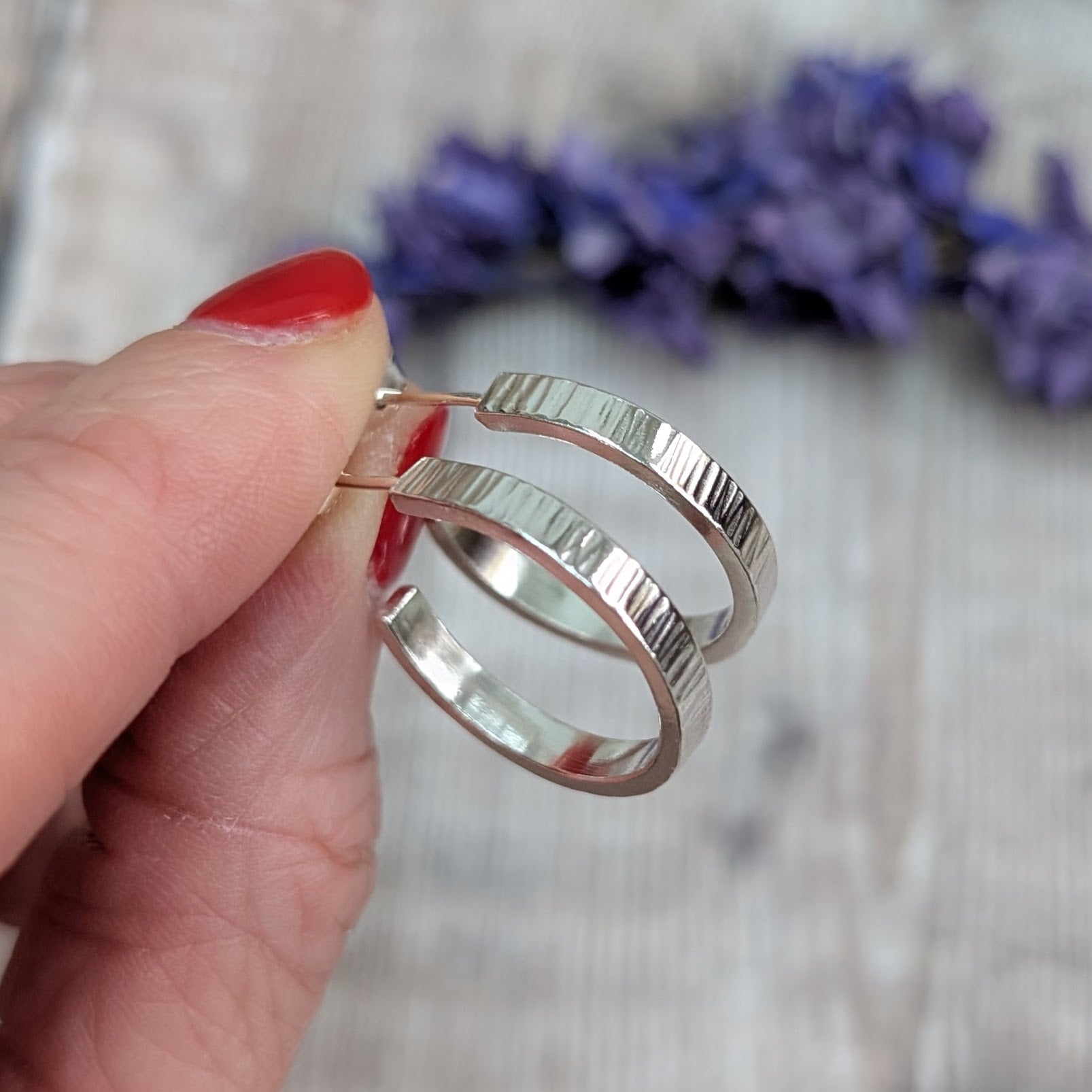 Hand holding a pair of Silver Bark Textured Hoop Earrings, showing the subtle bark-like hammered surface and curved shape against a blurred lavender background.