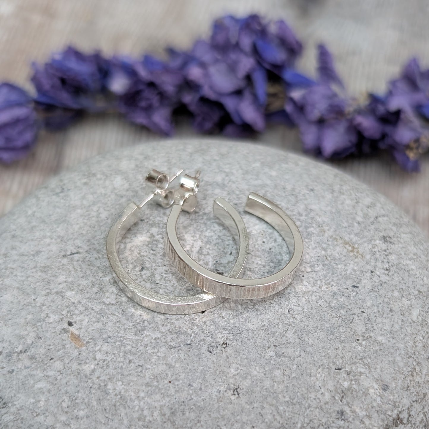 Silver Bark Textured Hoop Earrings resting on a smooth grey stone, with dried purple flowers in the background.