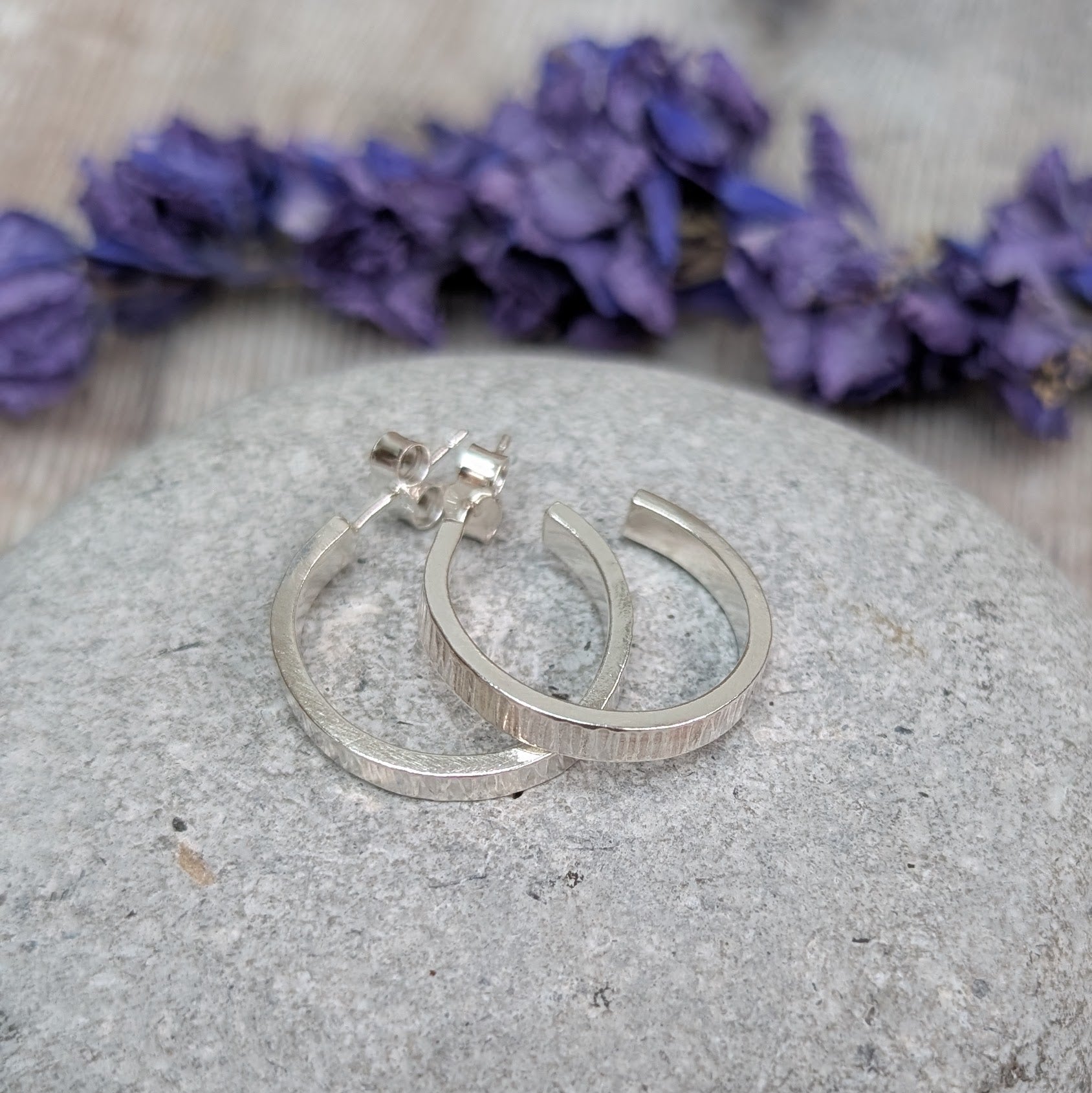 Silver Bark Textured Hoop Earrings resting on a smooth grey stone, with dried purple flowers in the background.