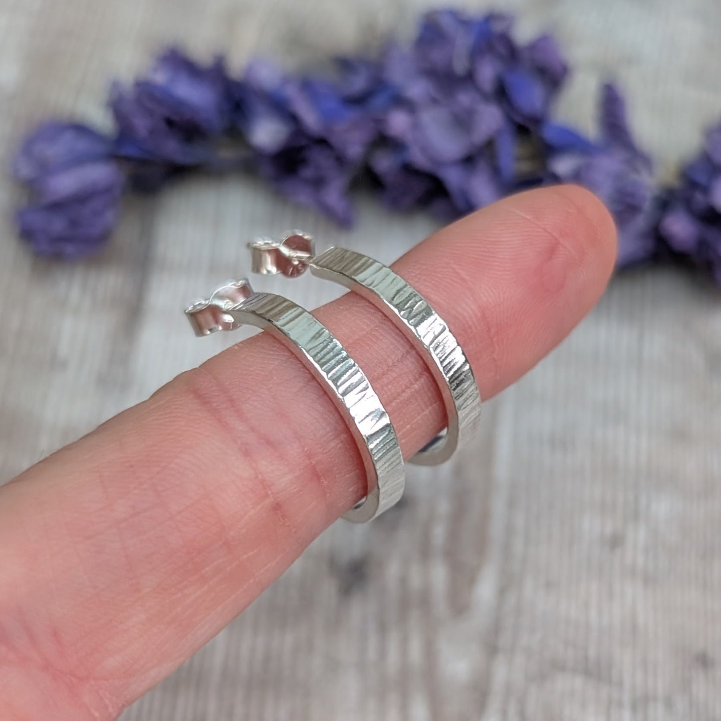 Pair of Silver Bark Textured Hoop Earrings balanced on a fingertip, showing the reflective bark-textured finish and curved form.