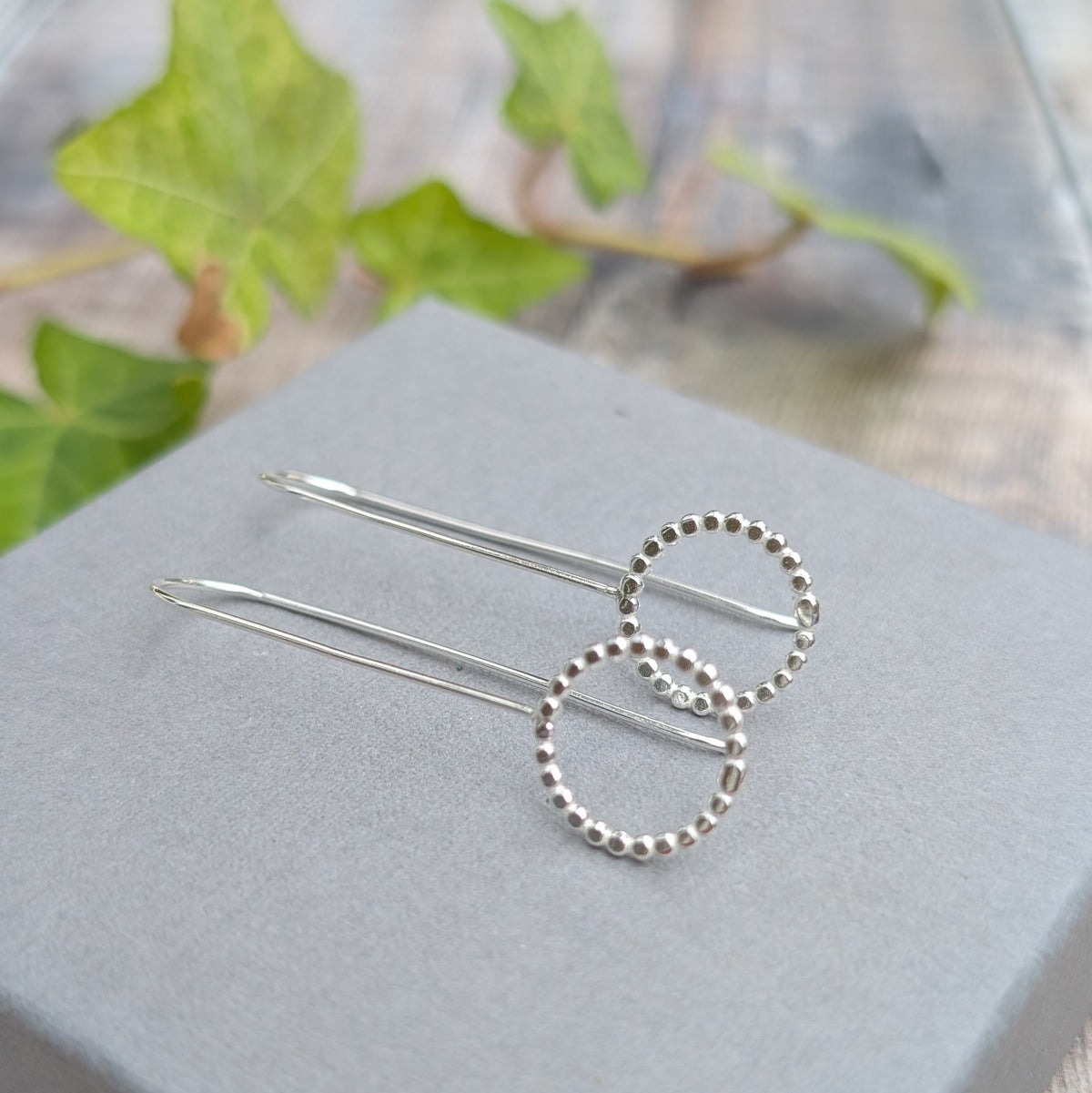 Silver beaded circle long earrings handmade from recycled Sterling Silver, displayed on a grey jewellery box with green ivy in the background.
