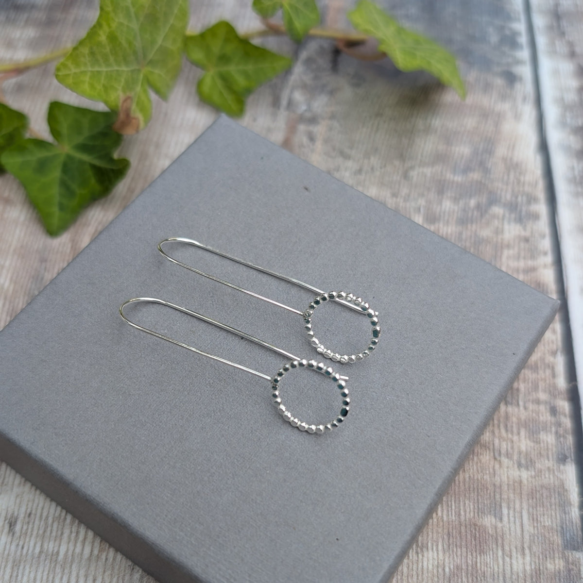 Silver beaded circle long earrings displayed on a grey jewellery box, showing the long hooks and textured open circles.