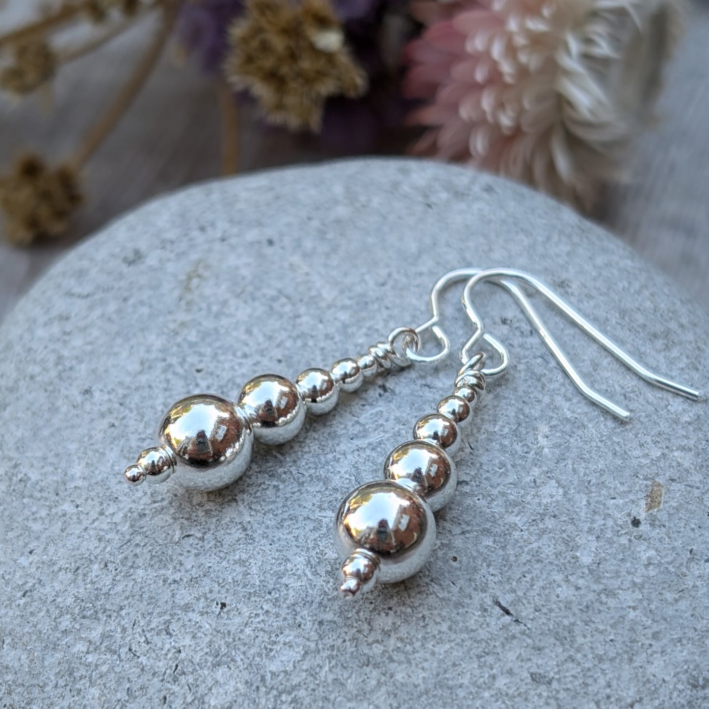 Close-up of a pair of handmade sterling silver drop earrings, featuring graduated round silver beads, resting on a smooth grey stone with dried flowers in the background.