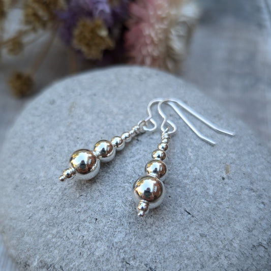 Close-up of a pair of handmade sterling silver drop earrings, featuring graduated round silver beads, resting on a smooth grey stone with dried flowers in the background.