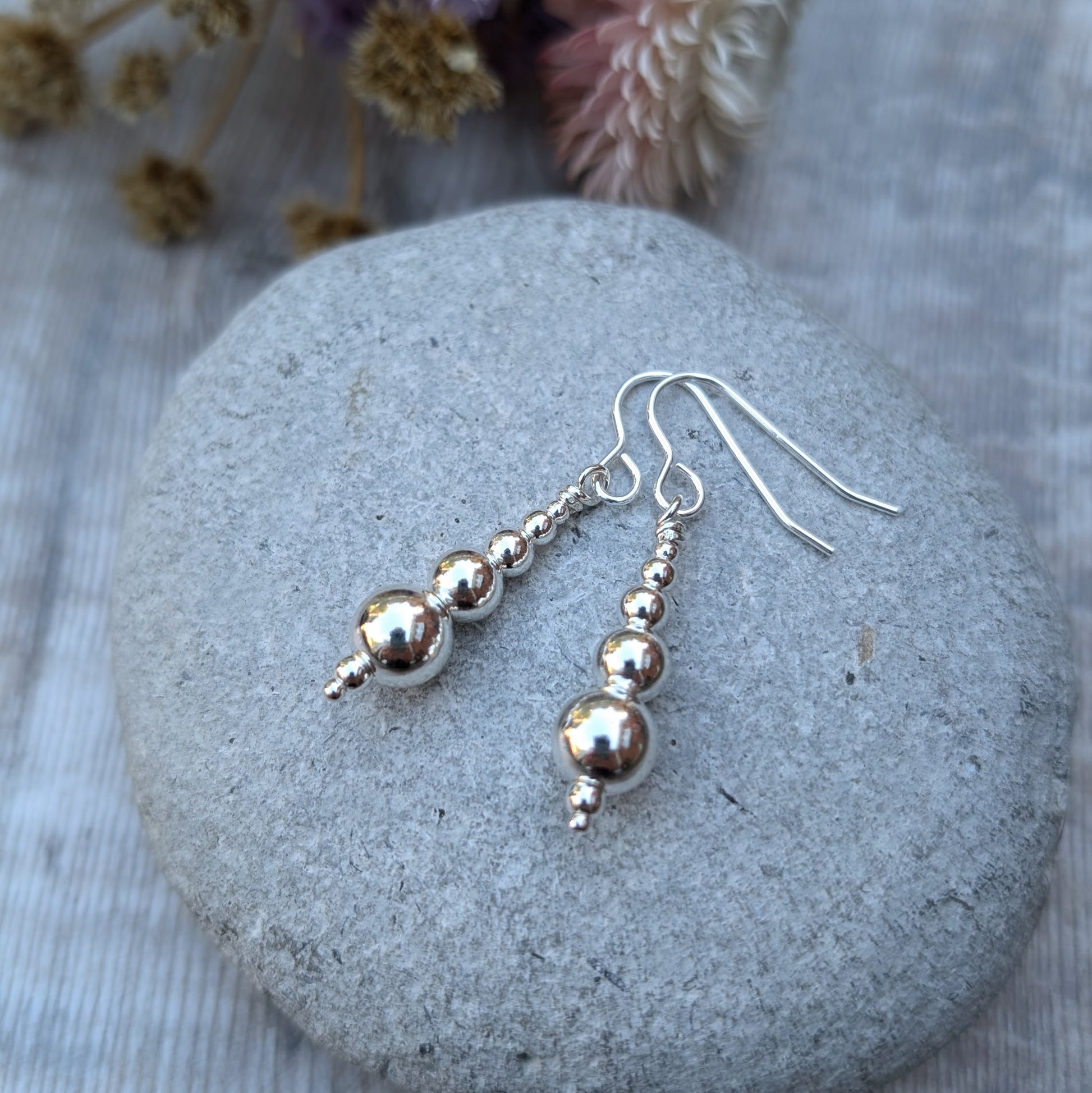 Close-up of a pair of handmade sterling silver drop earrings, featuring graduated round silver beads, resting on a smooth grey stone with dried flowers in the background.