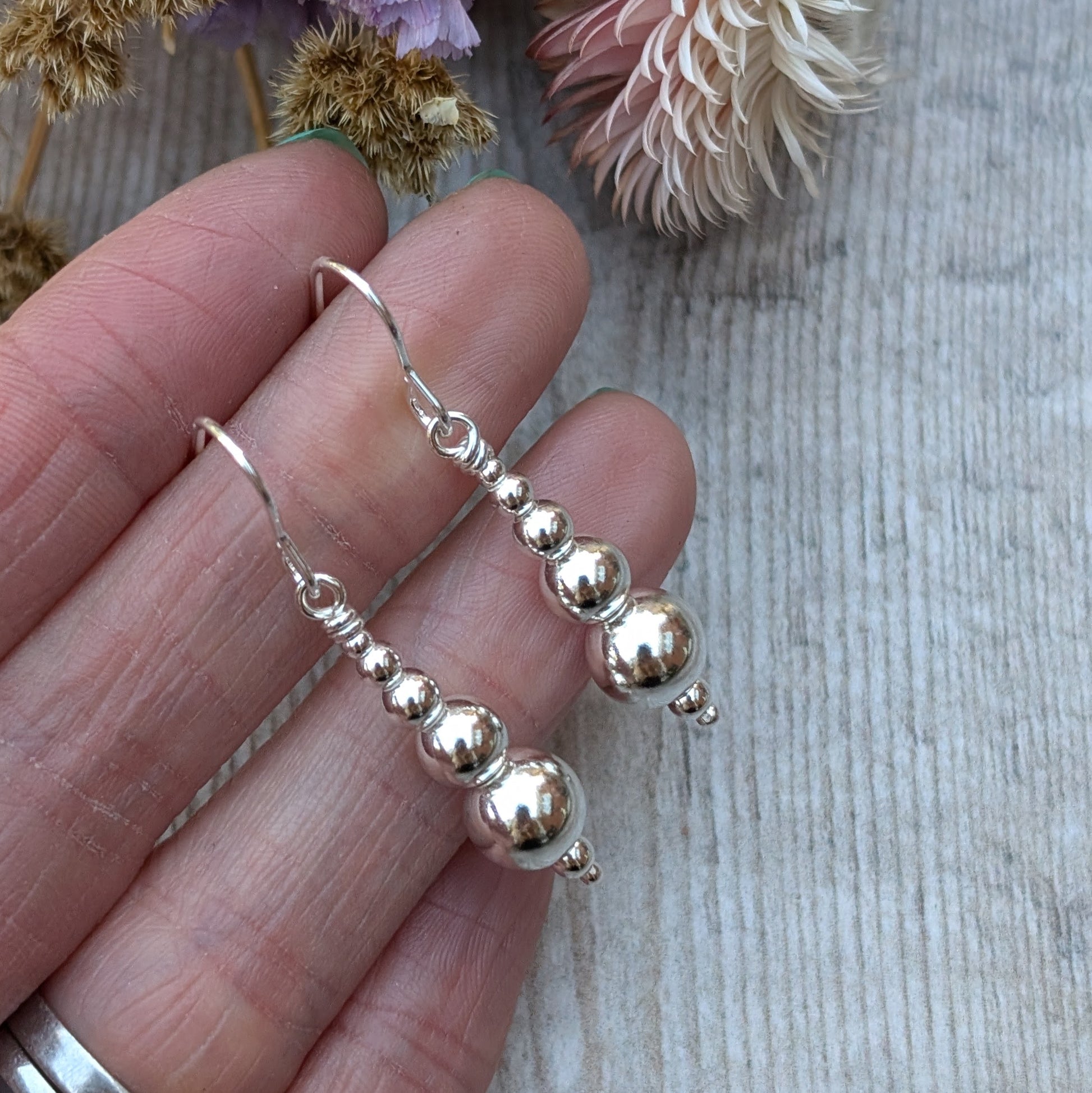 Close-up of a hand holding a pair of handmade sterling silver drop earrings, each featuring a series of polished round silver beads in descending sizes, with dried flowers in the background.