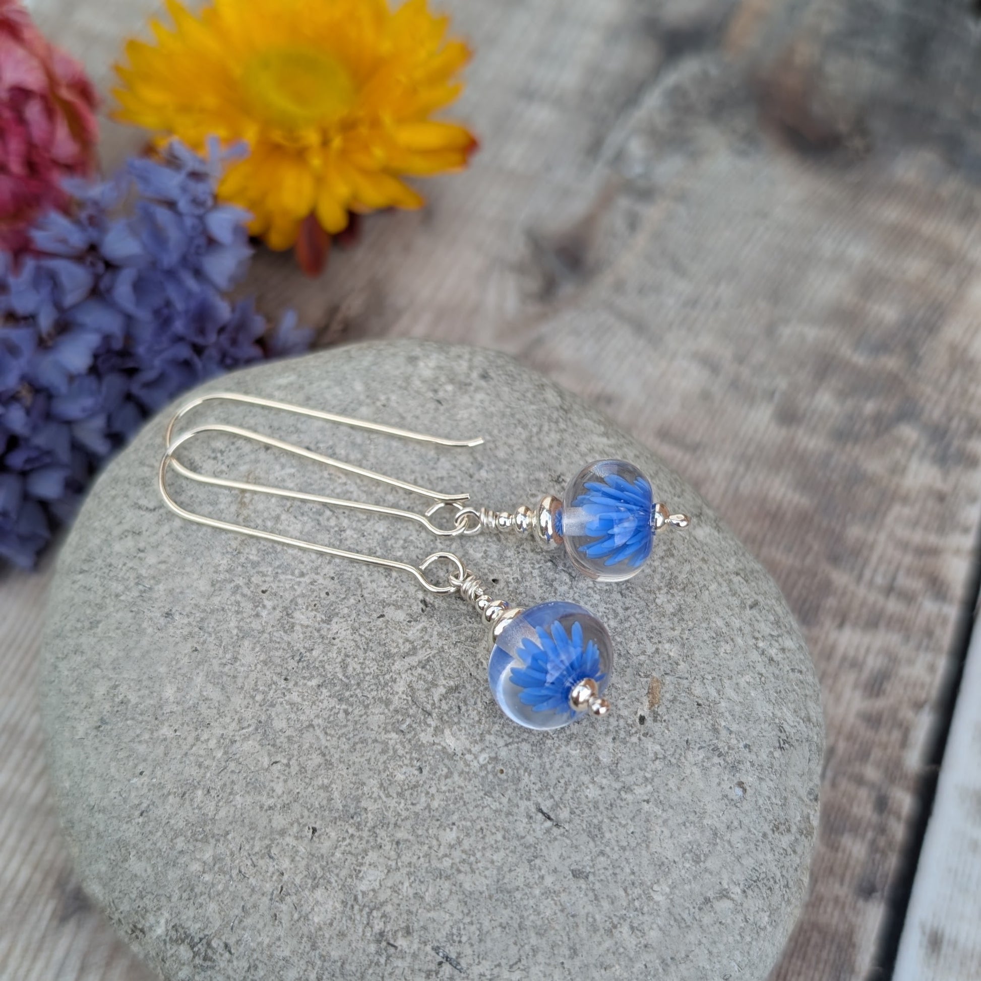 Silver Blue Lampwork Glass Earrings displayed on a smooth grey stone, featuring clear round glass beads with blue flower details and long silver ear wires.