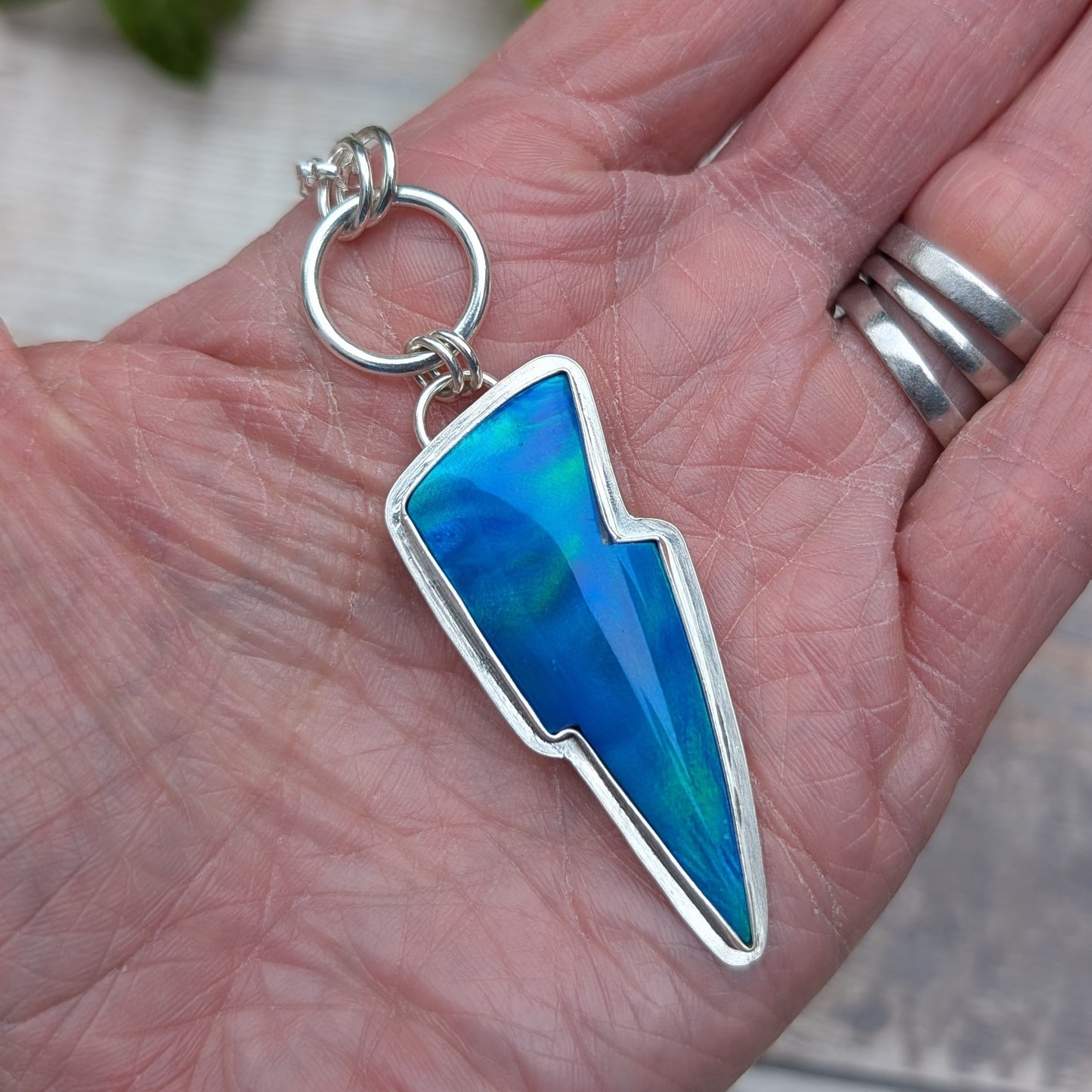 Silver Blue Aurora Opal Lightning Bolt Necklace shown in the palm of a hand, highlighting the blue and green colours of the Aurora Opal and the recycled sterling silver setting.