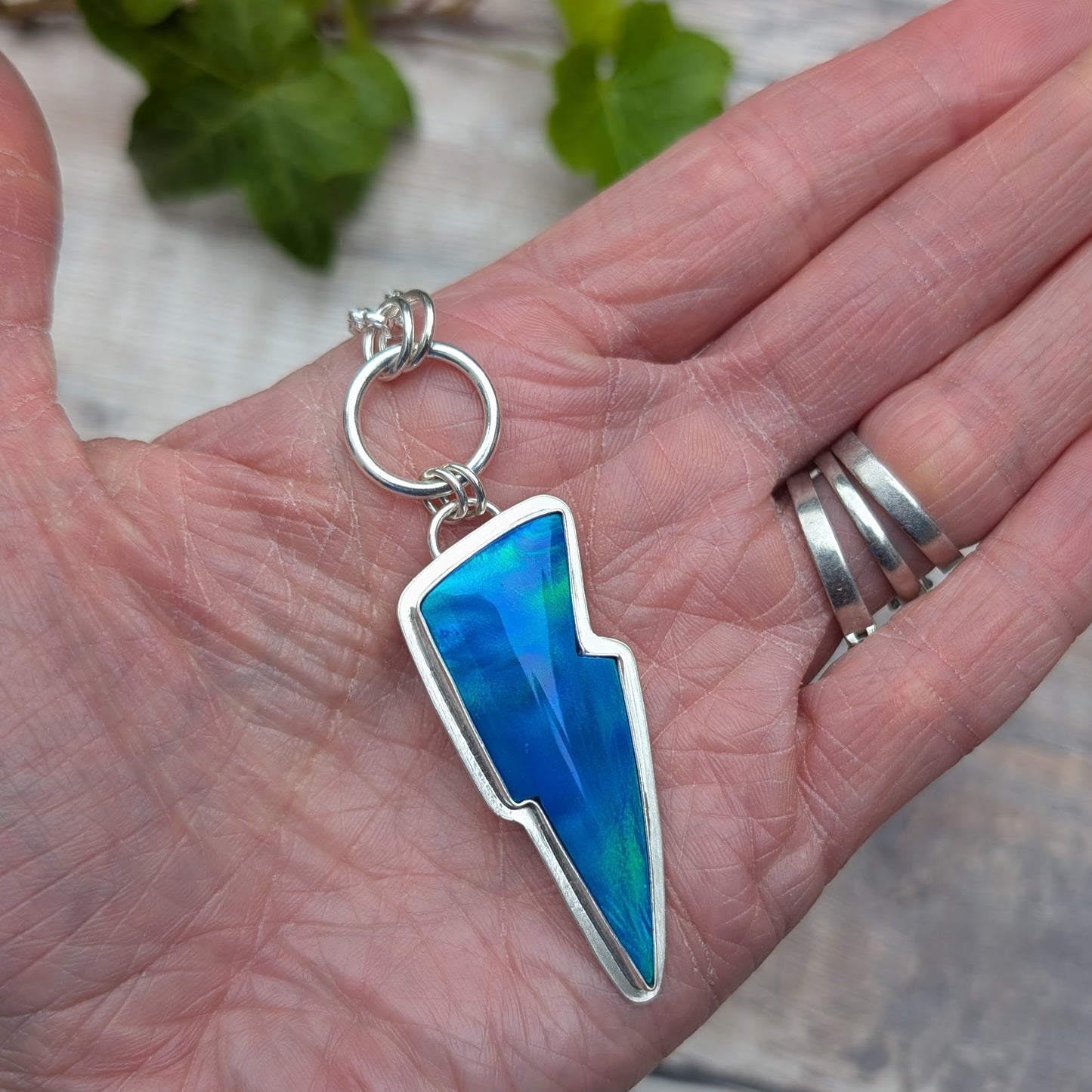 Silver Blue Aurora Opal Lightning Bolt Necklace resting in a hand, showing the lightning bolt shape, hand made sterling silver setting and colourful Aurora Opal stone.