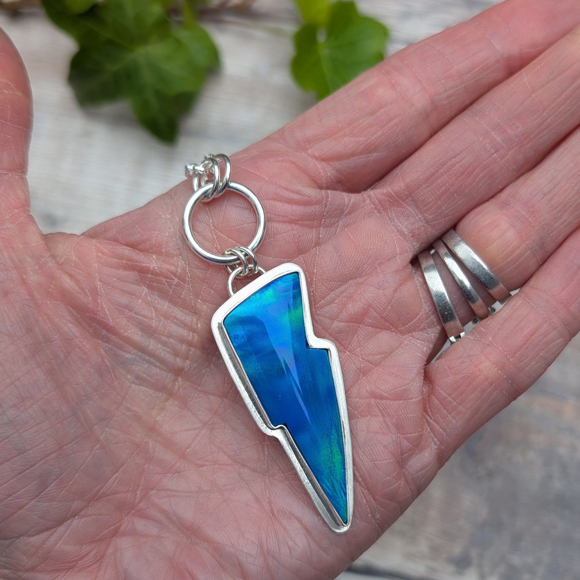 Silver Blue Aurora Opal Lightning Bolt Necklace resting in a hand, showing the lightning bolt shape, hand made sterling silver setting and colourful Aurora Opal stone.
