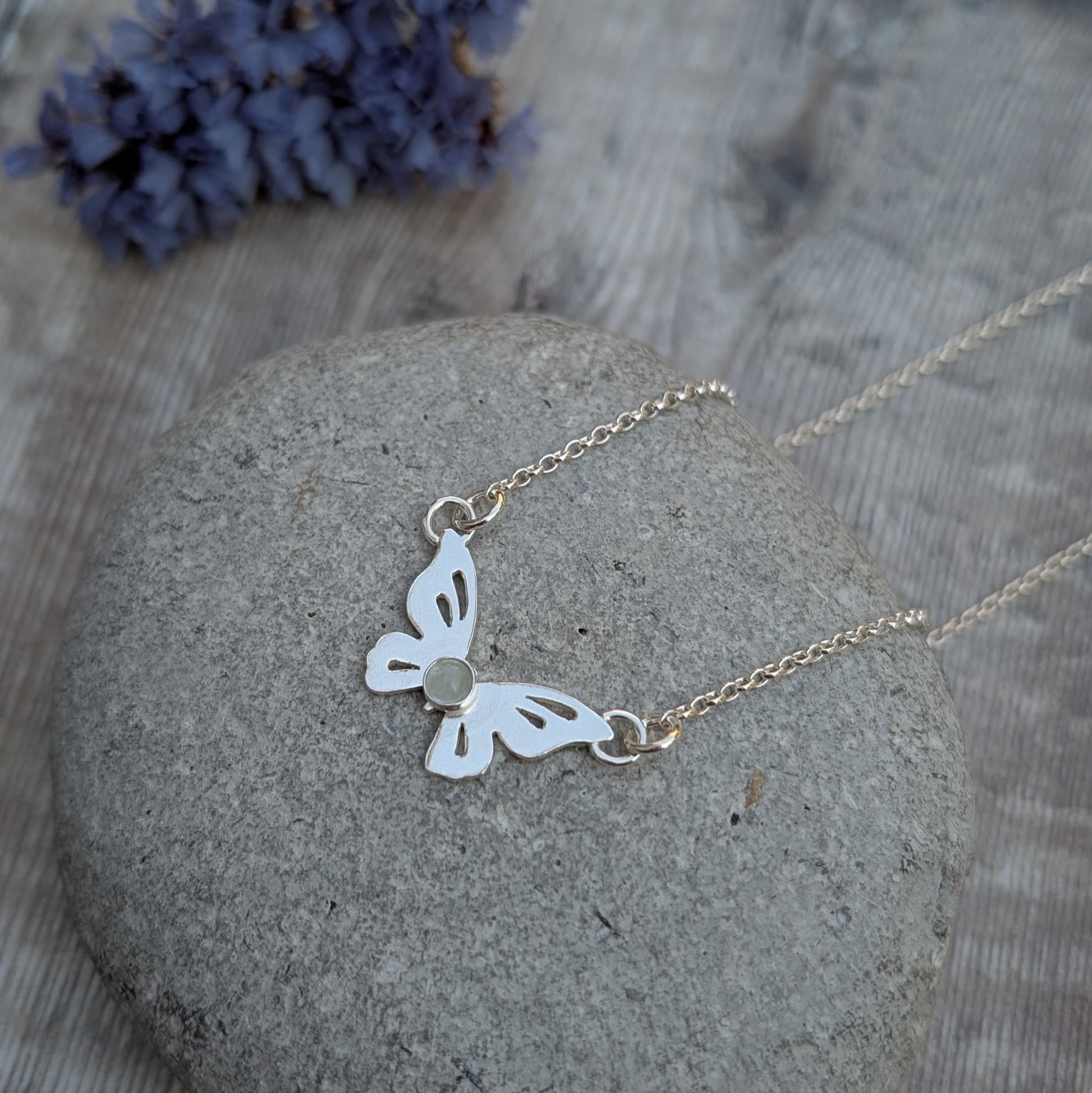 Silver Butterfly Necklace with Aquamarine resting on a smooth stone with soft purple flowers in the background