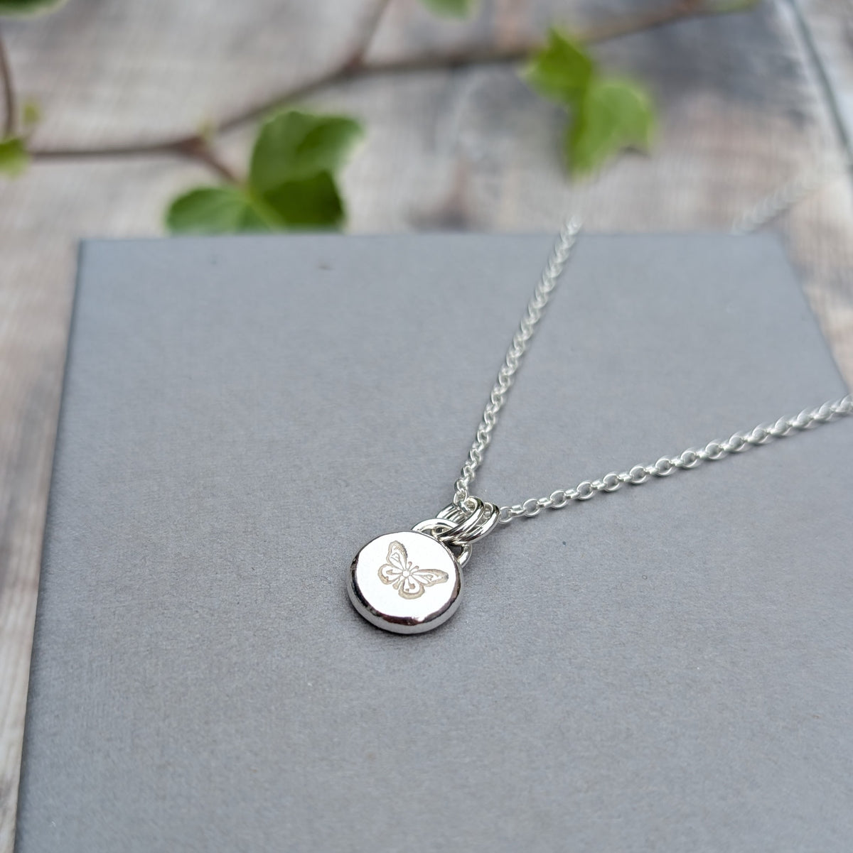 Close up of a handmade silver butterfly pebble necklace with a stamped butterfly design, displayed on a grey background.