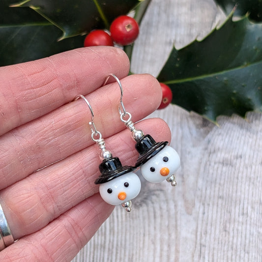 Close-up of a hand holding the Silver Christmas Snowman Lampwork Glass Earrings, showing the white glass snowman beads with black hats, tiny black eyes, and orange noses.