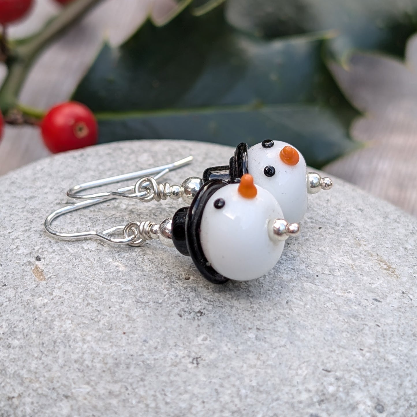 The Silver Christmas Snowman Lampwork Glass Earrings resting on a stone, showing the handmade snowman beads with black hats and orange noses from a side angle.