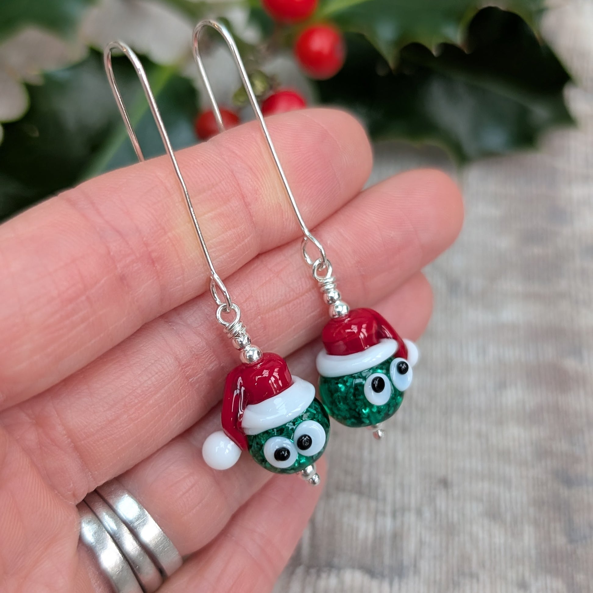 Close-up of a hand holding the Silver Christmas Sparkly Sprout Lampwork Glass Earrings, highlighting the glittery green sprout beads, white detailing, and bright red Santa hats.