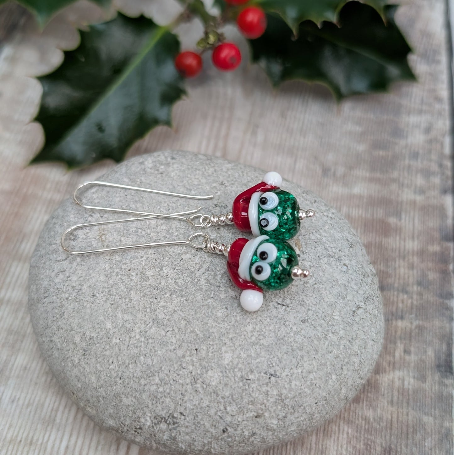 Close-up of a pair of Silver Christmas Sparkly Sprout Lampwork Glass Earrings resting on a stone, showing the green sparkly sprout beads wearing red and white Santa hats on long sterling silver ear wires.