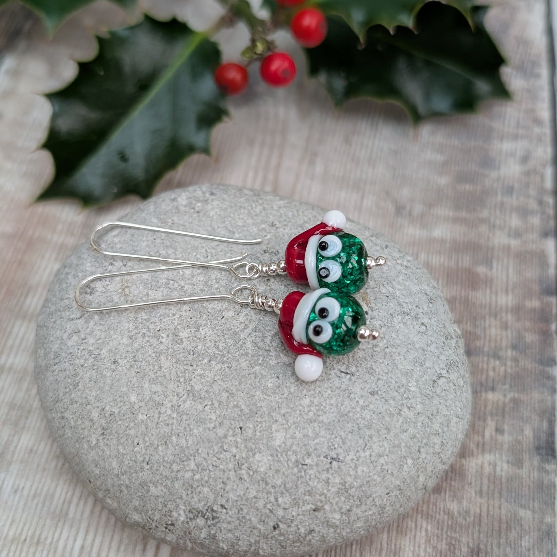 Close-up of a pair of Silver Christmas Sparkly Sprout Lampwork Glass Earrings resting on a stone, showing the green sparkly sprout beads wearing red and white Santa hats on long sterling silver ear wires.
