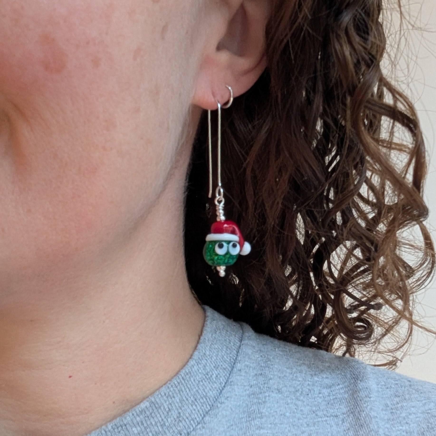 A person wearing one of the Silver Christmas Sparkly Sprout Lampwork Glass Earrings, showing the long silver ear wire and the sparkly green sprout bead with a tiny Santa hat.