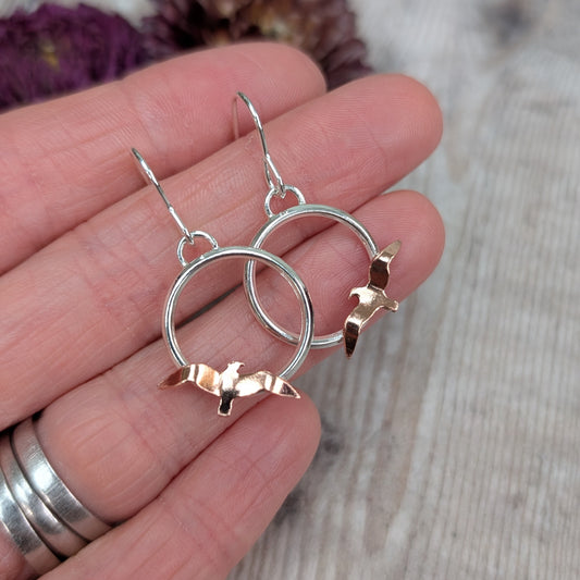 A pair of Silver Circle Earrings with Copper Birds held gently between fingers, showing the small hand-cut copper birds and smooth silver circles with a soft wooden background behind.